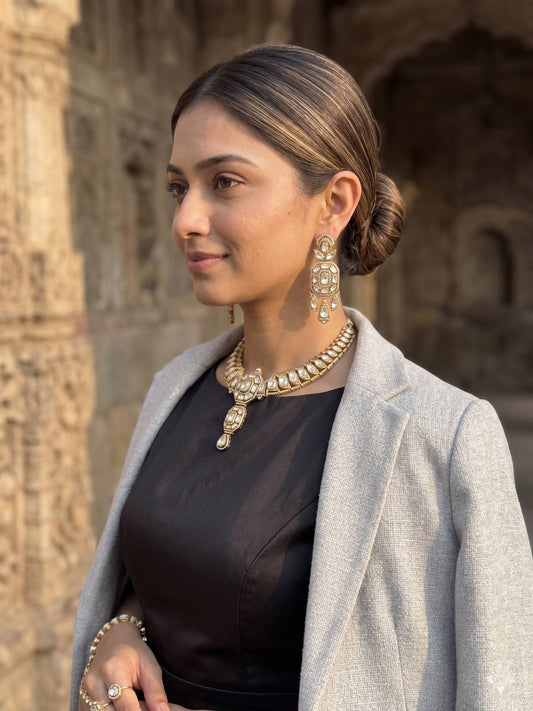 Woman wearing a black outfit with a gray coat and gold jewelry, standing in front of an architectural background.
