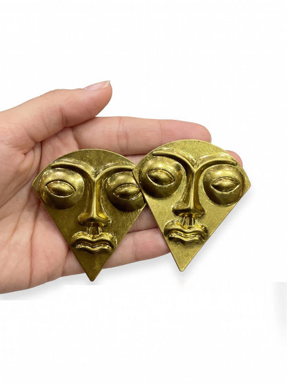 Gold face-shaped earrings held in a hand against a white background