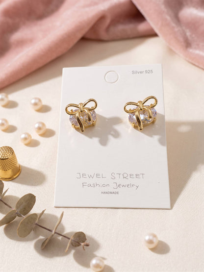 Gold bow-shaped earrings on a 'Jewel Street' branded card with pearls and leaves around.