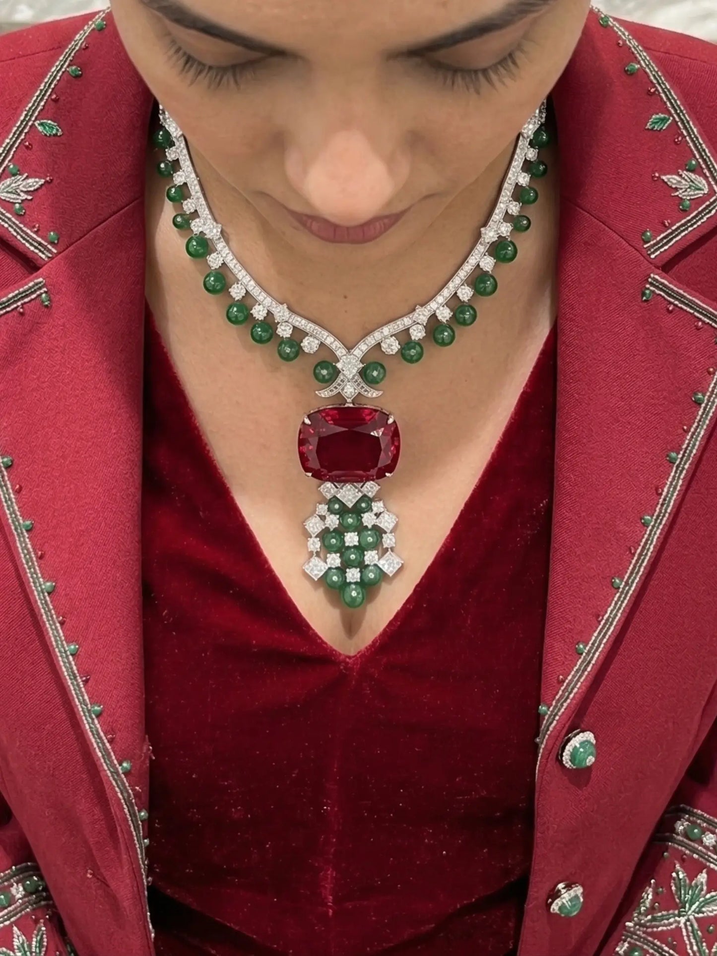 Person wearing a luxurious emerald and diamond necklace with a red gemstone on a red outfit.