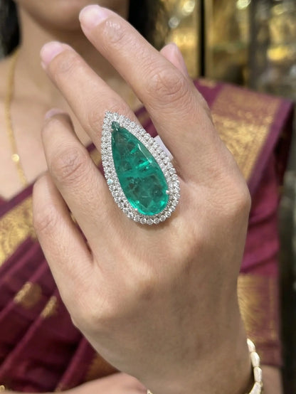 A close-up photograph of a woman wearing a large, elegant pear-shaped green cubic zirconia ring on her middle finger.