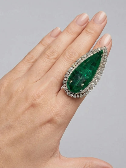 Hand wearing a ring with a large green gemstone and diamond accents on a gray background