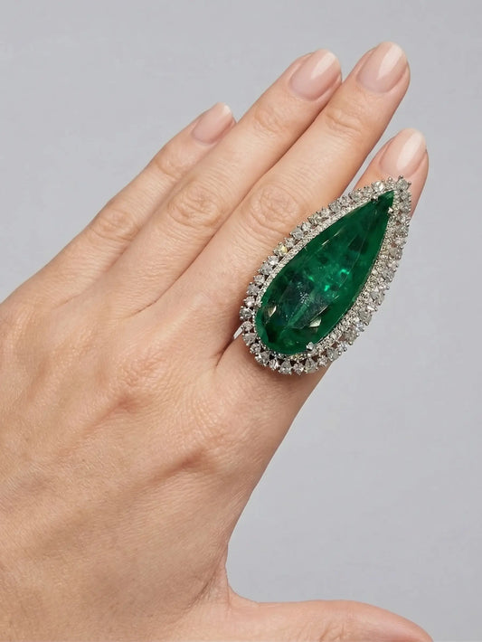 Hand wearing a ring with a large green gemstone and diamond accents on a gray background