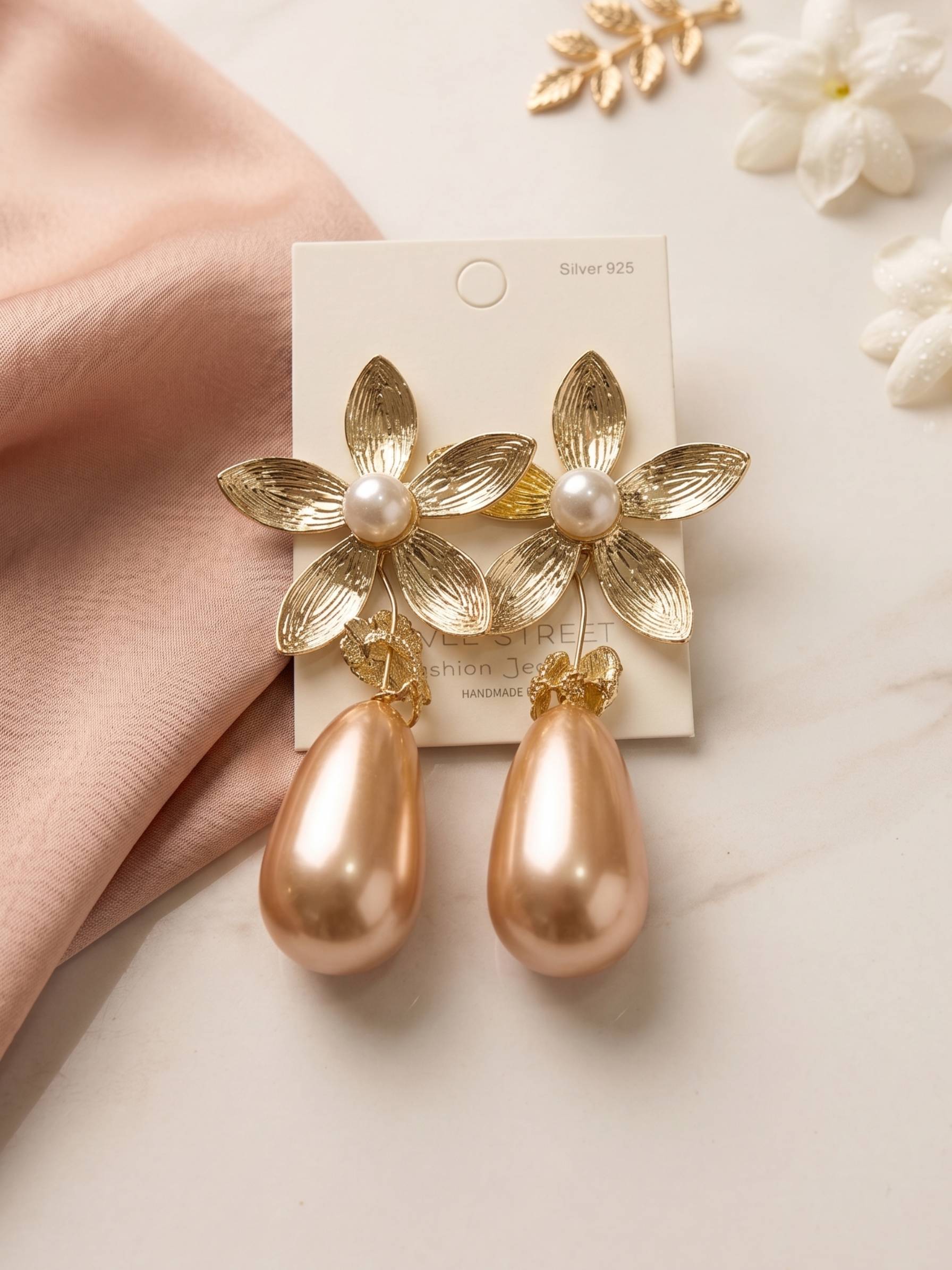 Gold floral earrings with pearl accents on a card, held by a hand with a soft background.