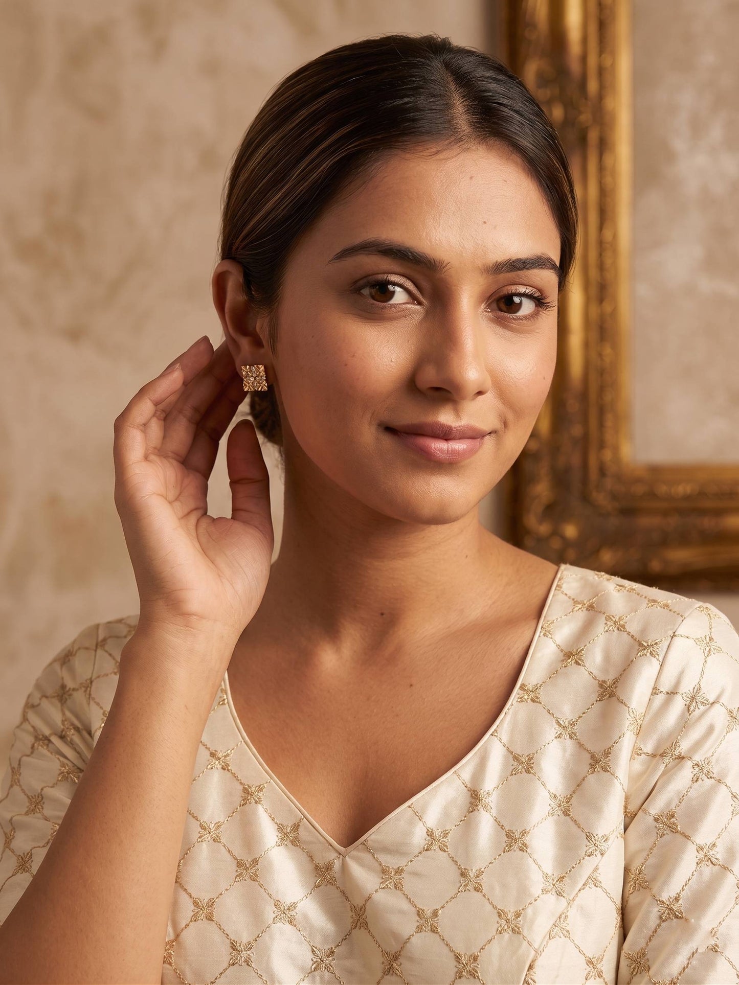 Woman wearing a beige patterned top with a gold earring, standing against a warm-toned wall.