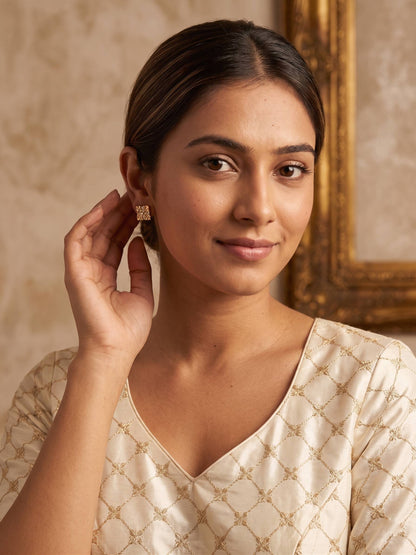 Woman wearing a beige patterned top with a gold earring, standing against a warm-toned wall.