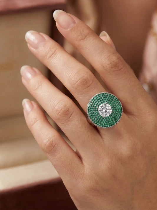 Close-up of a hand wearing a green ring with a diamond on a neutral background