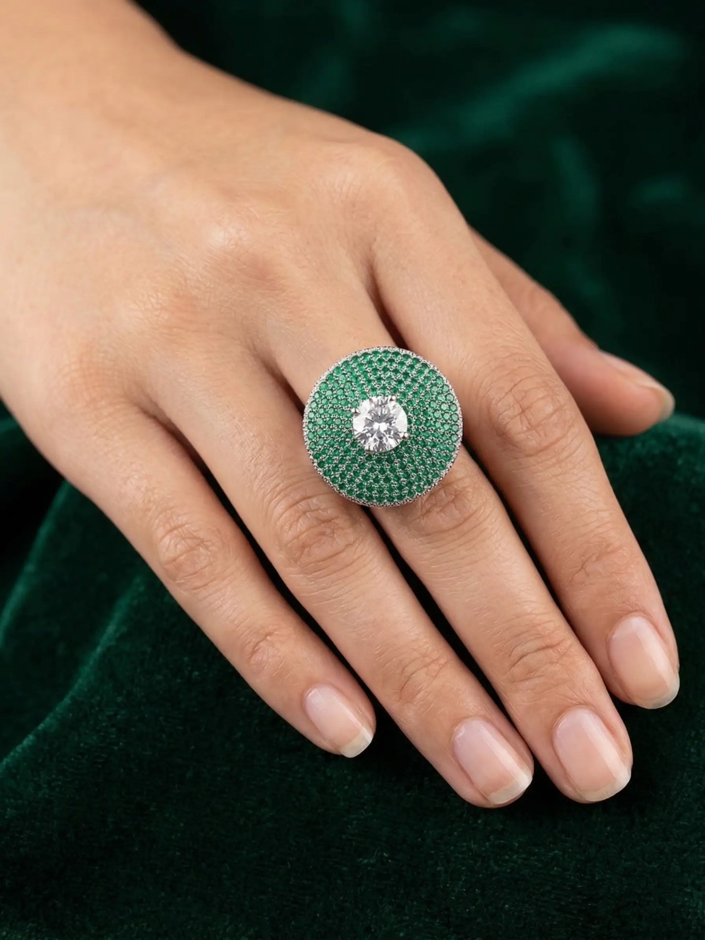 Close-up of a hand wearing a green gemstone ring with a diamond center on a dark green background.