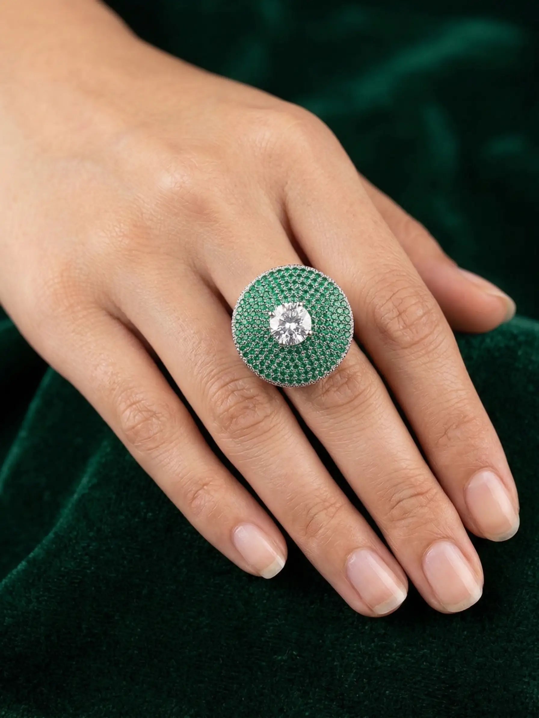 Close-up of a hand wearing a green gemstone ring with a diamond center on a dark green background.