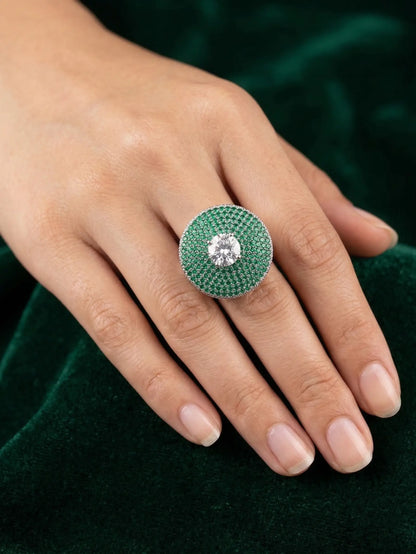 Close-up of a hand wearing a green gemstone ring with a diamond center on a dark green background.