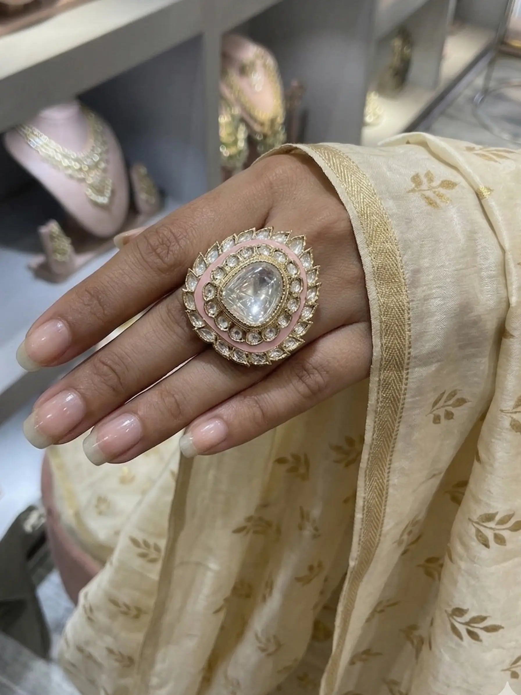 Hand wearing a decorative ring with a clear gemstone, set against a blurred jewelry store background.
