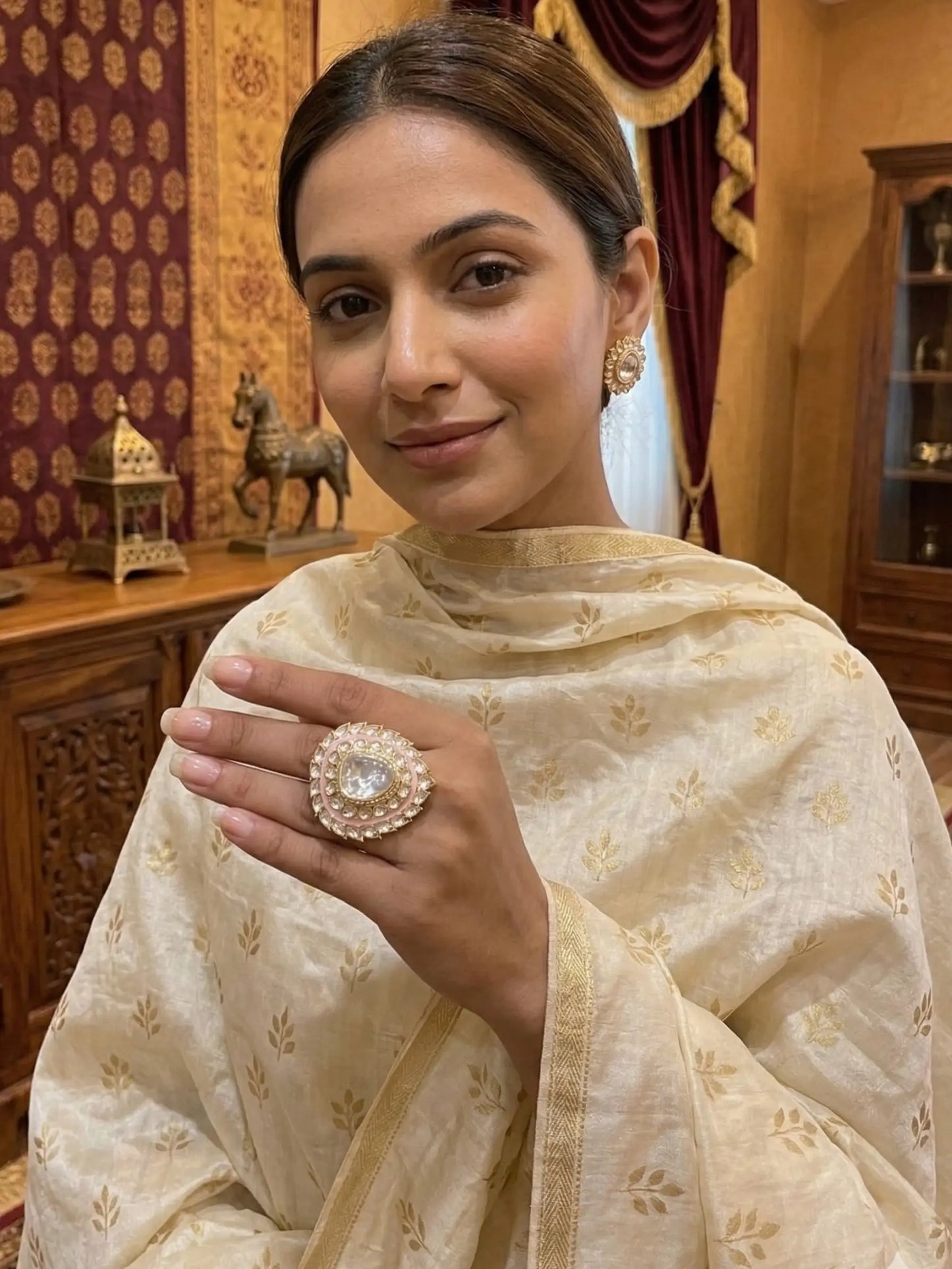 Woman in a cream-colored traditional outfit holding a decorative ring indoors.