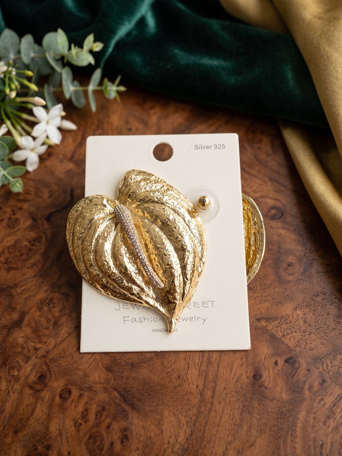 Gold leaf-shaped earring on a card with greenery and fabric in the background