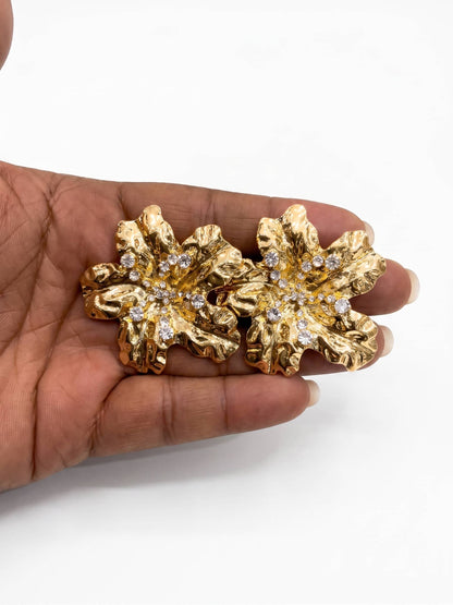 Gold floral earrings with embedded stones held in a hand against a white background