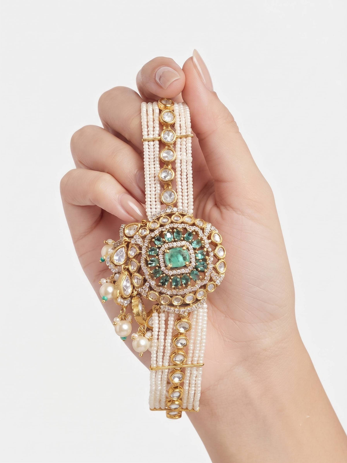 A hand holding a traditional Indian Kundan bracelet with a central green stone, gold-toned beaded bands, and clear rhinestone embellishments, set against a plain light gray background.