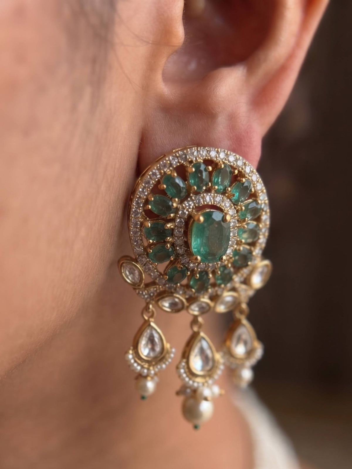A close-up of a person's ear wearing a large, ornate gold earring decorated with green emerald-cut stones and small clear pavé accents, with three dangling teardrop charms.