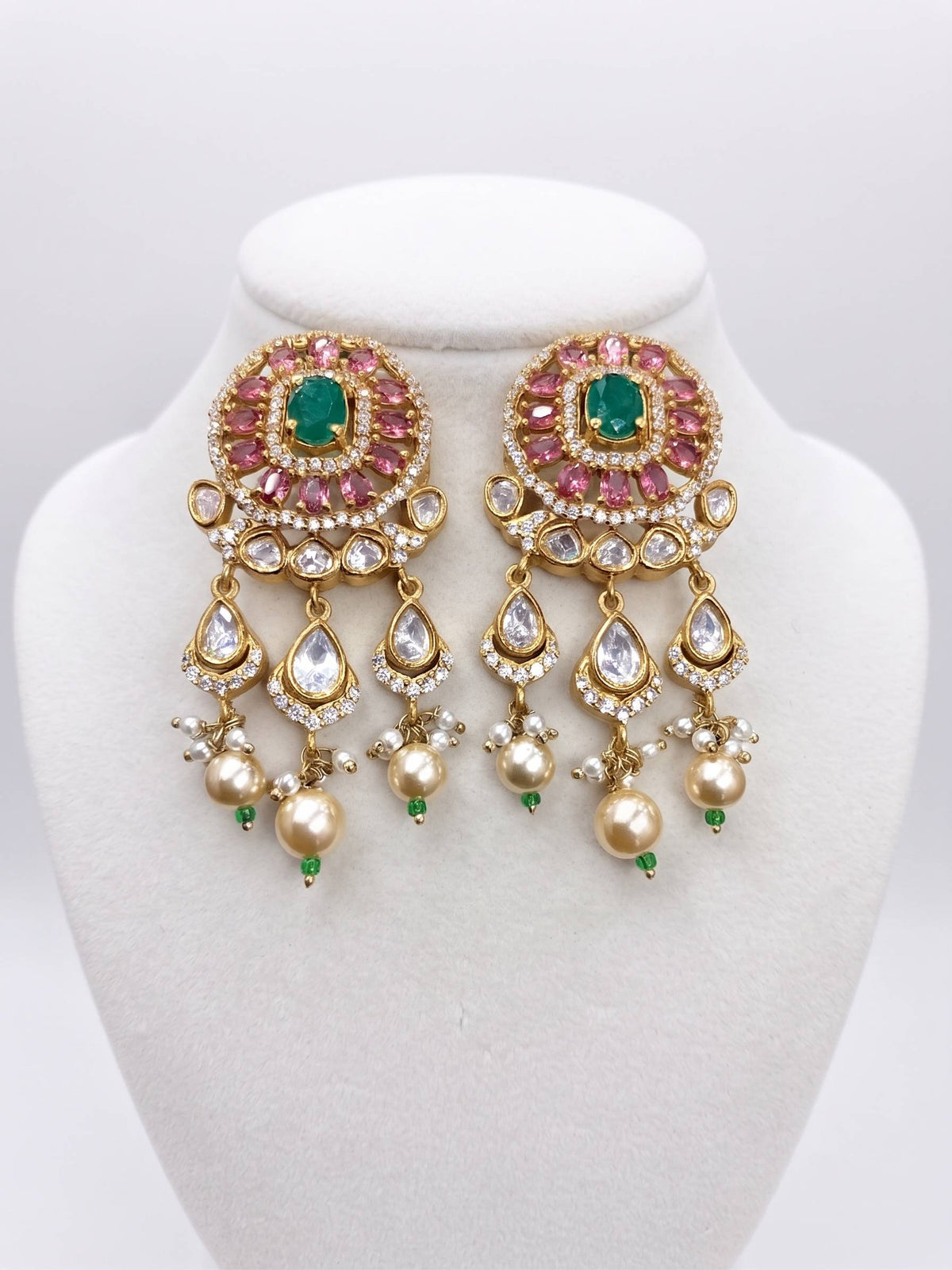 A pair of ornate gold-toned Indian chandbali earrings with green emerald-cut stones, pink round stones, clear teardrop crystals, and gold pearl-like drops, displayed on a white jewelry bust.