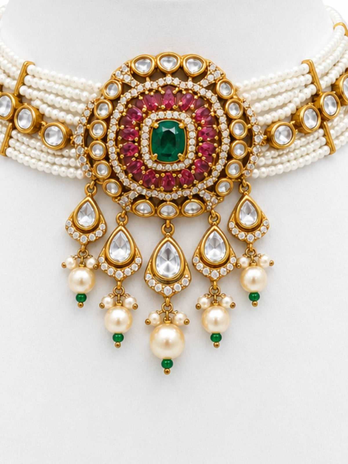 An ornate Indian style choker necklace featuring a gold-plated centerpiece with a central green emerald, surrounded by pinkish-red stones and clear stones, with multiple strands of white pearls and dangling teardrop charms.