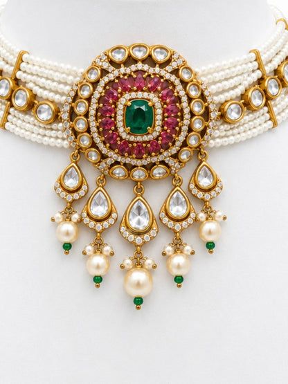 An ornate Indian style choker necklace featuring a gold-plated centerpiece with a central green emerald, surrounded by pinkish-red stones and clear stones, with multiple strands of white pearls and dangling teardrop charms.