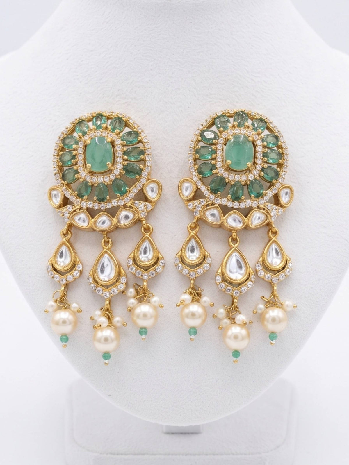 A pair of ornate gold-plated Indian chandelier earrings with green stones, white faux pearls, and clear crystals, displayed on a white jewelry bust.