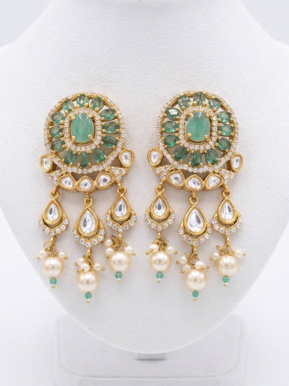 A pair of ornate gold-plated Indian chandelier earrings with green stones, white faux pearls, and clear crystals, displayed on a white jewelry bust.