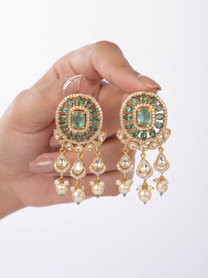 A hand holding a pair of ornate gold and green gemstone dangle earrings with pearl-like drops against a clean white background.