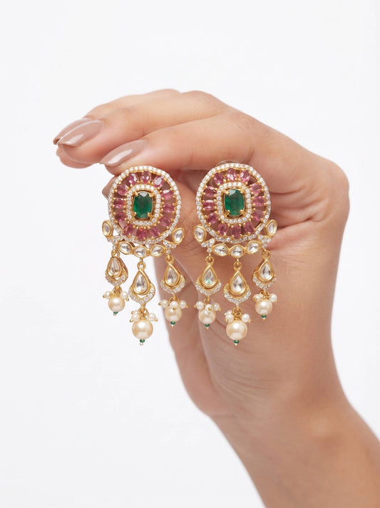 A close-up of a hand holding a pair of ornate gold-toned statement earrings with green rectangular center stones, pink stones around them, and dangling pearl-like teardrop and bead charms, against a plain white background.
