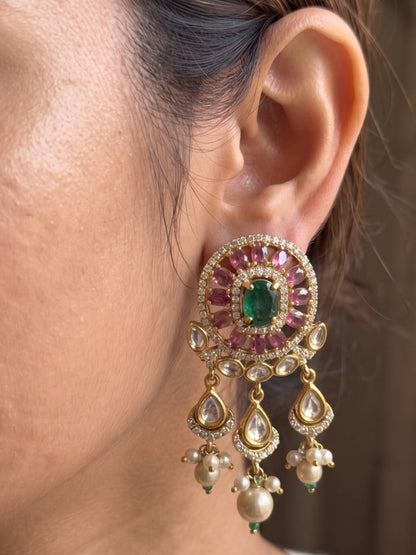 A close-up of a person's ear wearing an ornate gold-toned traditional Indian earring with a central green stone, surrounded by pink stones and clear stones, with dangling pearl-like and teardrop elements.