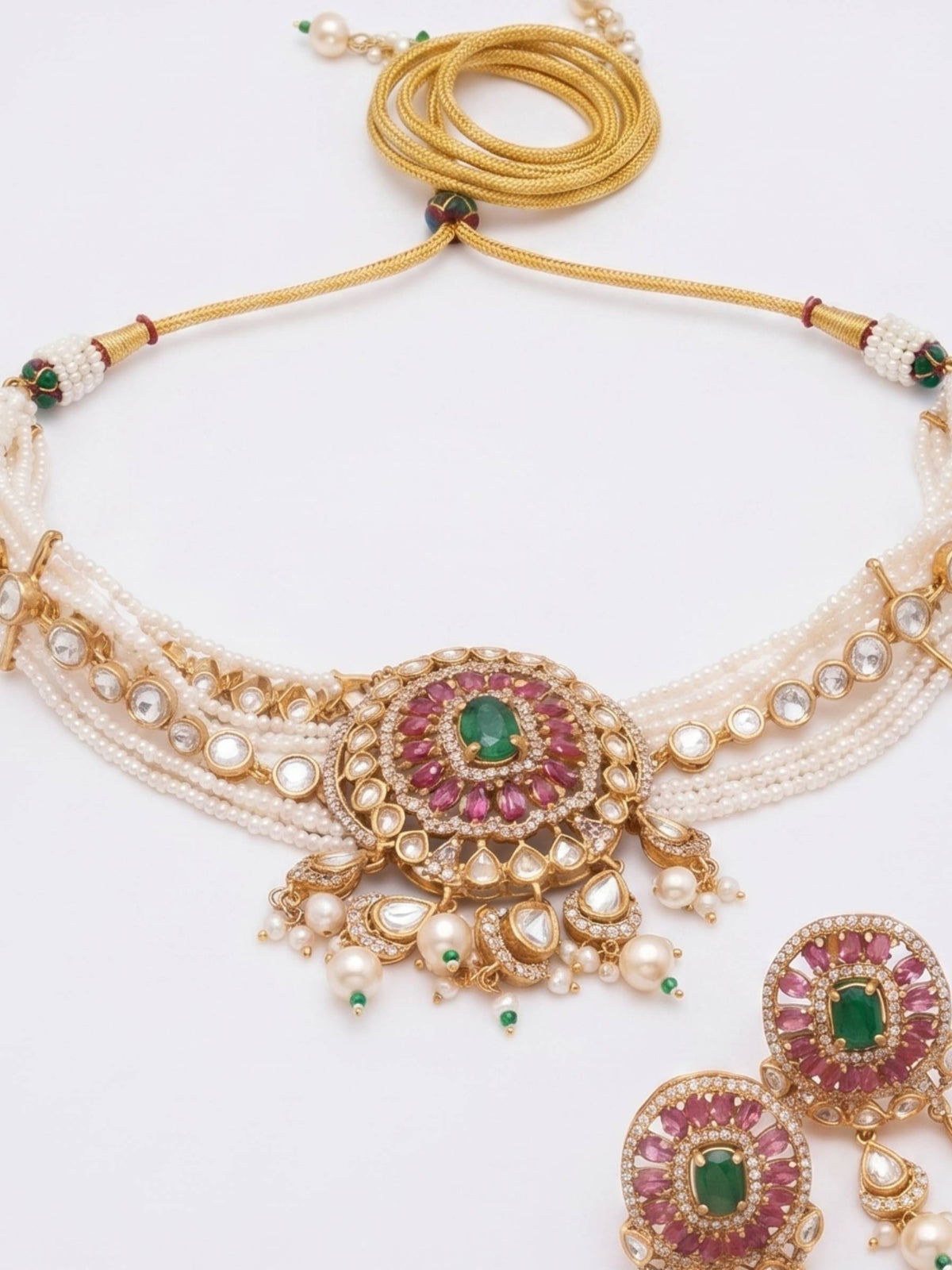 An ornate traditional Indian jewelry set featuring a multi-strand necklace with a central gold and green stone medallion, matching drop earrings, and a gold chain strap, displayed on a clean white background.