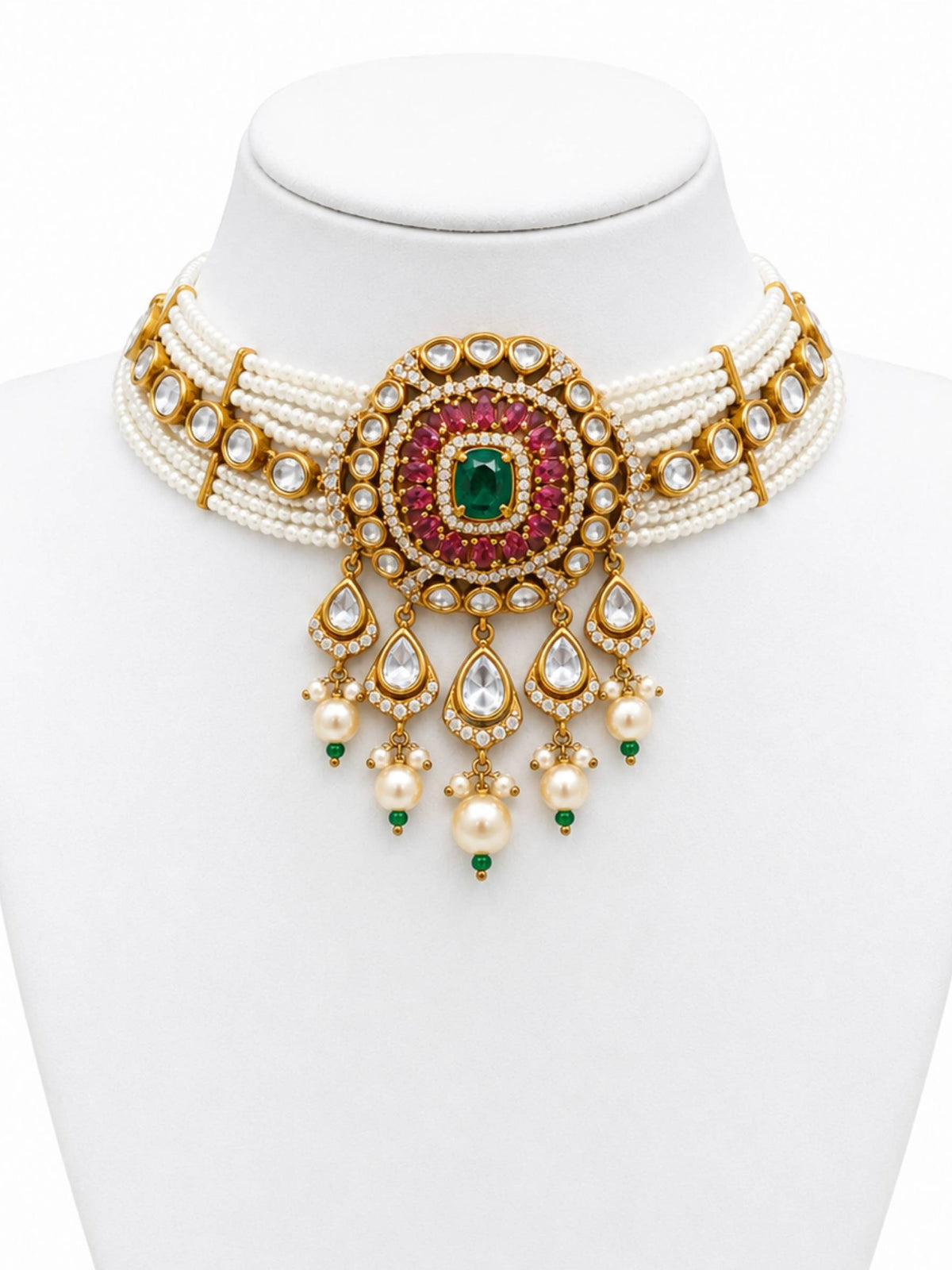 An ornate Indian style choker necklace displayed on a white mannequin bust. The necklace features a multi-strand white pearl base with a central gold-toned medallion decorated with a green emerald-cut stone, pink stones, and clear stones, with teardrop-shaped dangling charms.