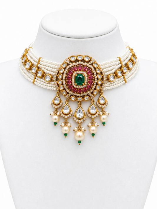 An ornate Indian style choker necklace displayed on a white mannequin bust. The necklace features a multi-strand white pearl base with a central gold-toned medallion decorated with a green emerald-cut stone, pink stones, and clear stones, with teardrop-shaped dangling charms.