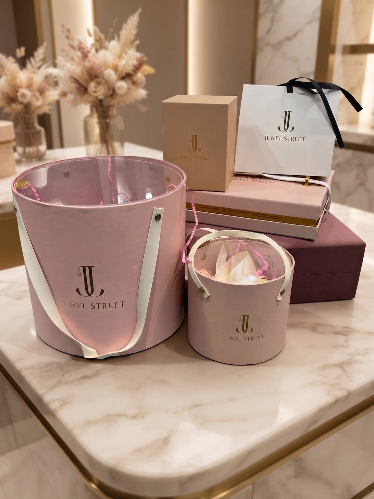 Pink jewelry gift packaging set from Jewel Street, including a large pink bucket bag, a smaller pink cylindrical bag, a white branded shopping bag, and matching boxes, arranged on a marble countertop.