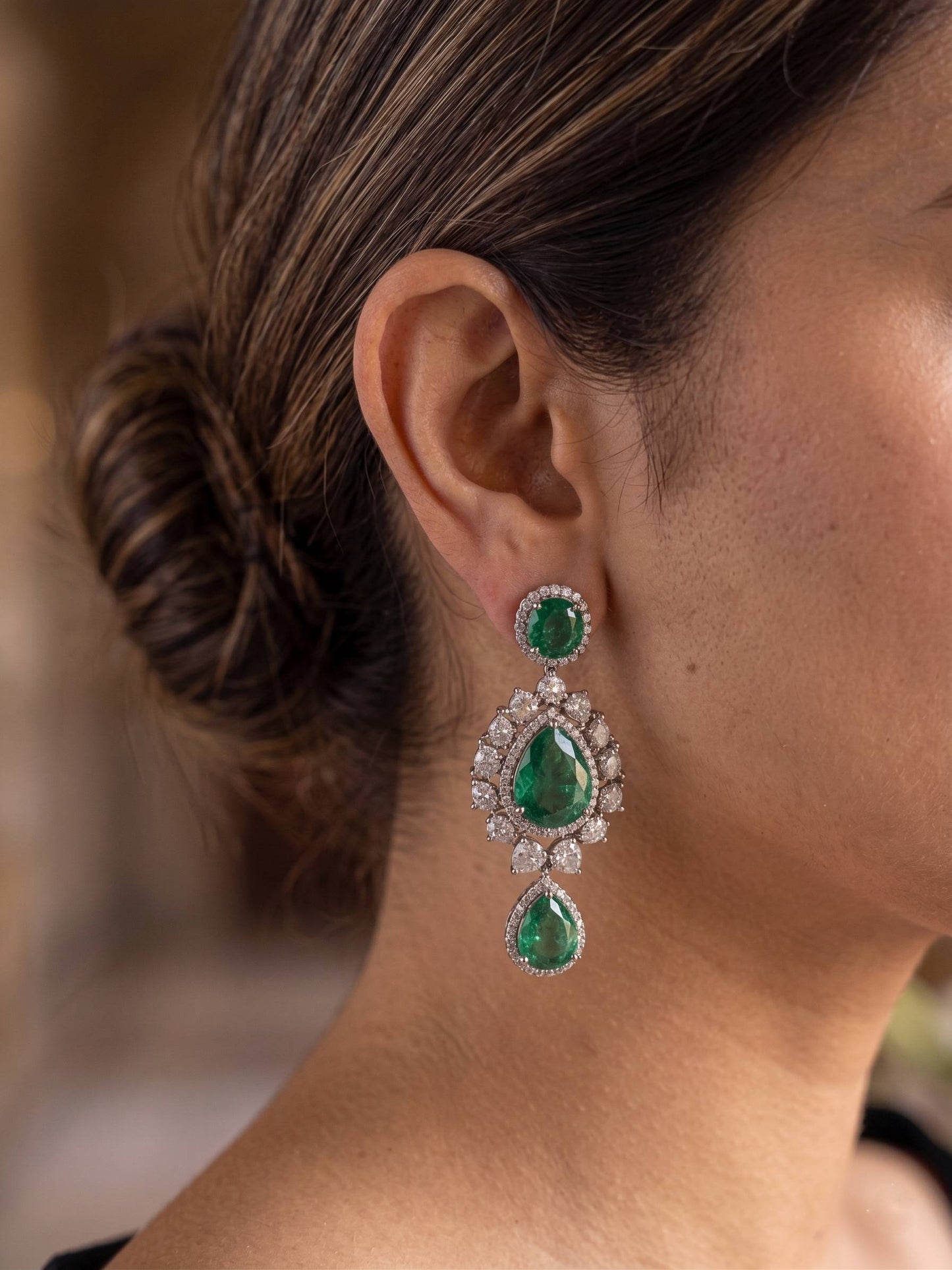 Close-up of a person wearing emerald and diamond earrings with a blurred background