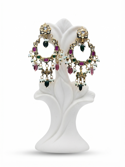 A pair of ornate gold-toned statement earrings with dangling floral and circular motifs, embellished with pink, green, and white stones, displayed on a white sculptural stand against a white background.