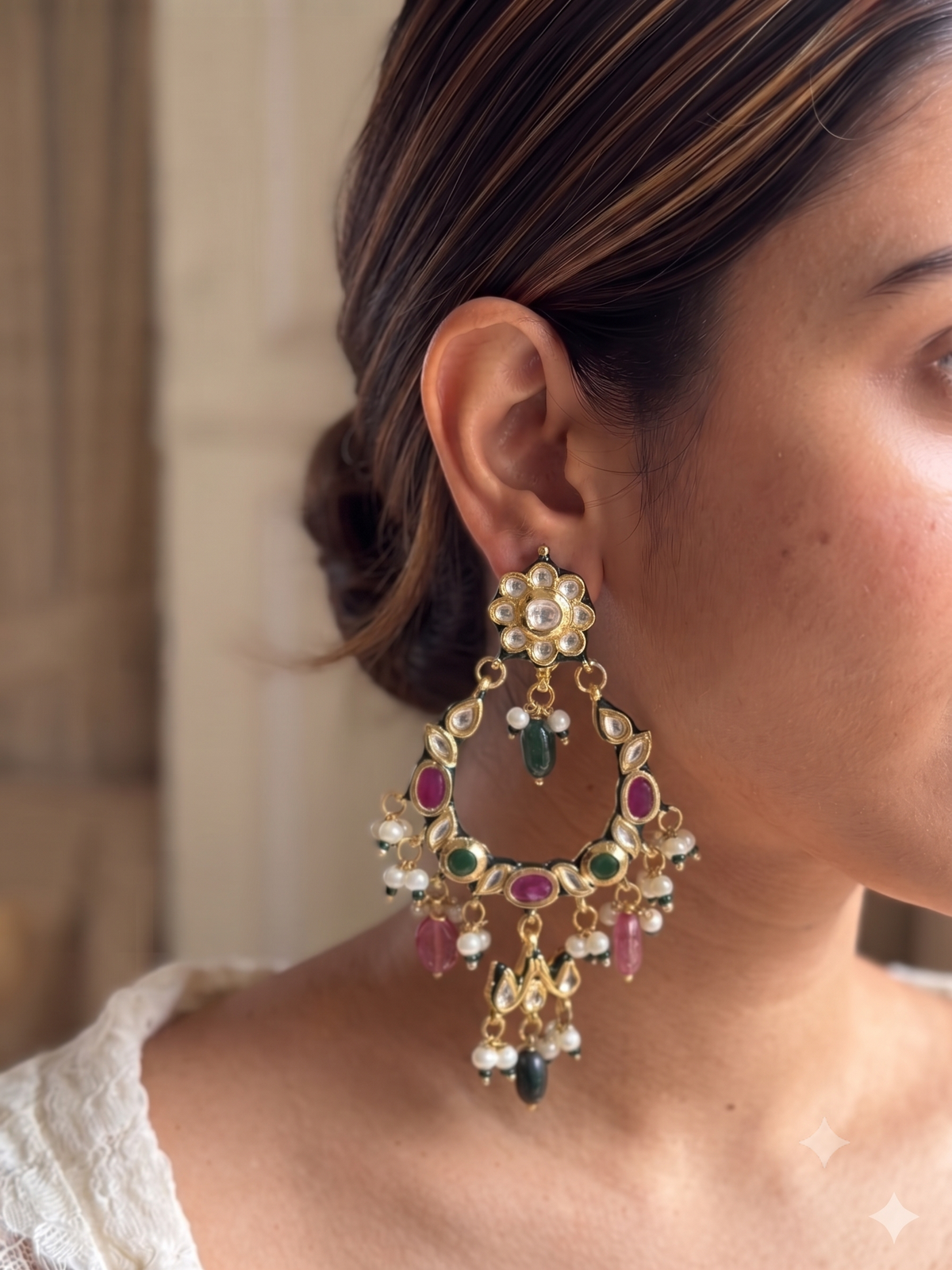 A close-up of a woman wearing a large, ornate gold-toned dangle earring decorated with green, pink, and white stones.