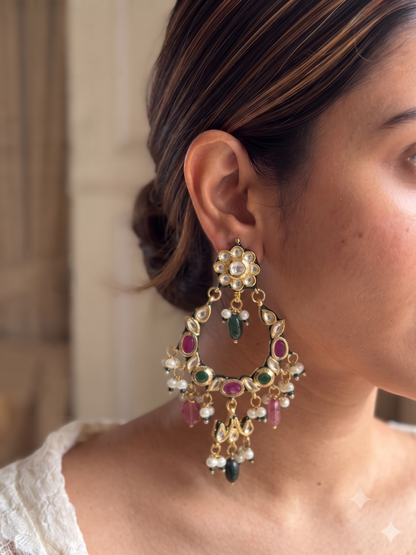 A close-up of a woman wearing a large, ornate gold-toned dangle earring decorated with green, pink, and white stones.