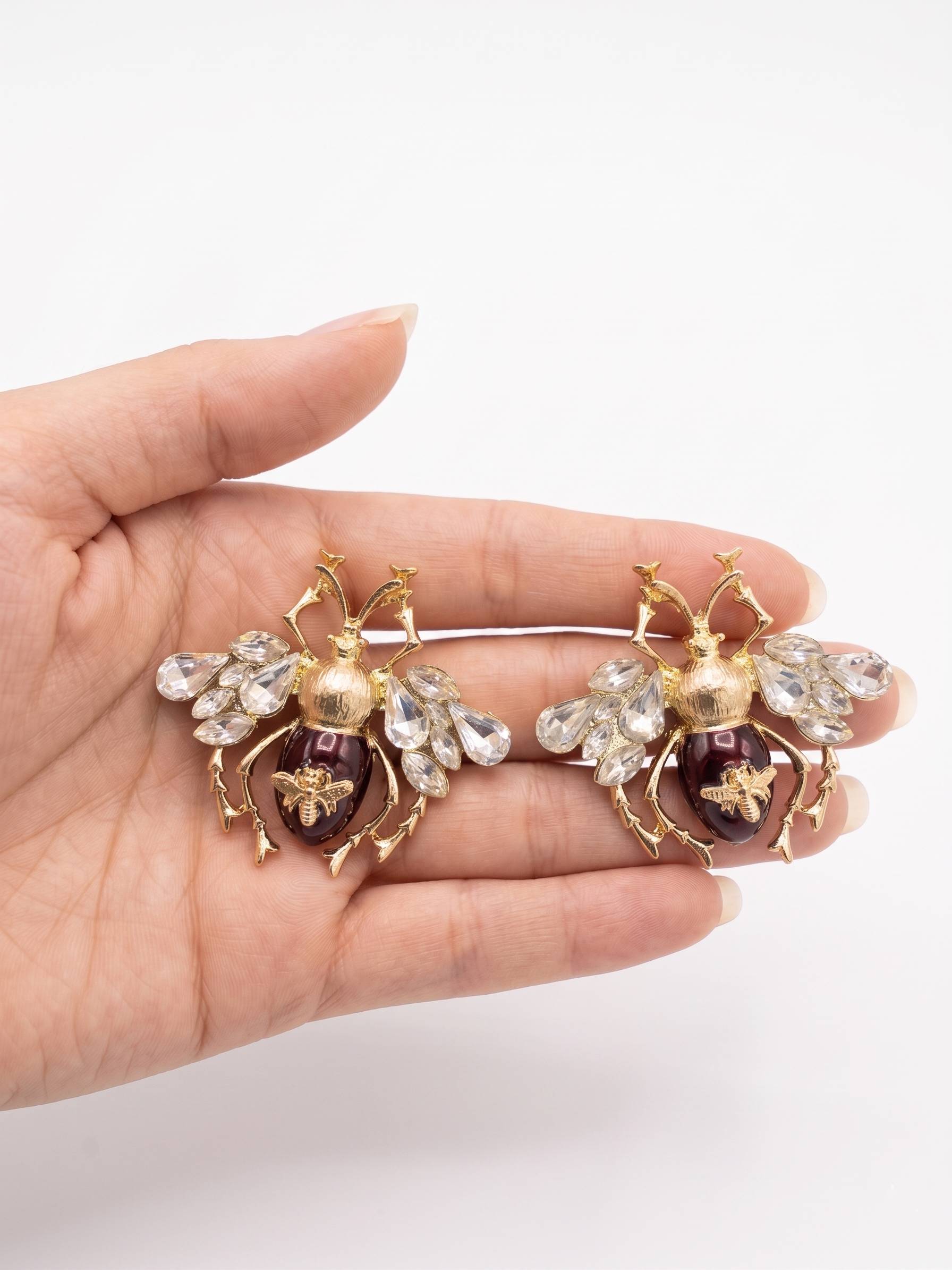 Hand holding two decorative bee-shaped brooches with gemstones on a white background