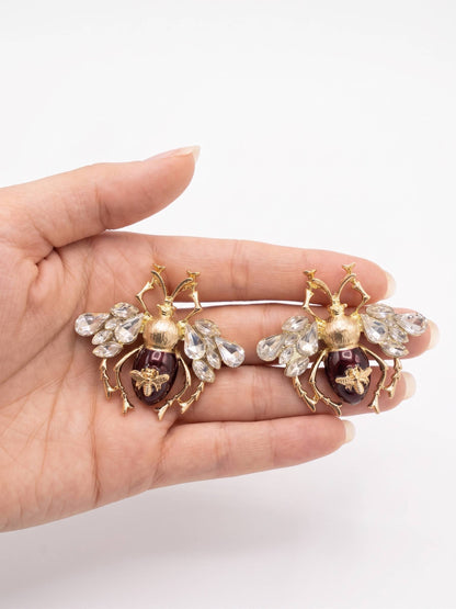 Hand holding two decorative bee-shaped brooches with gemstones on a white background