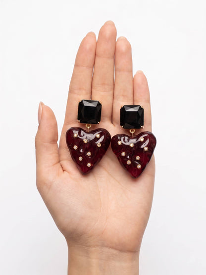 Heart-shaped earrings with black square stones held in a hand against a white background