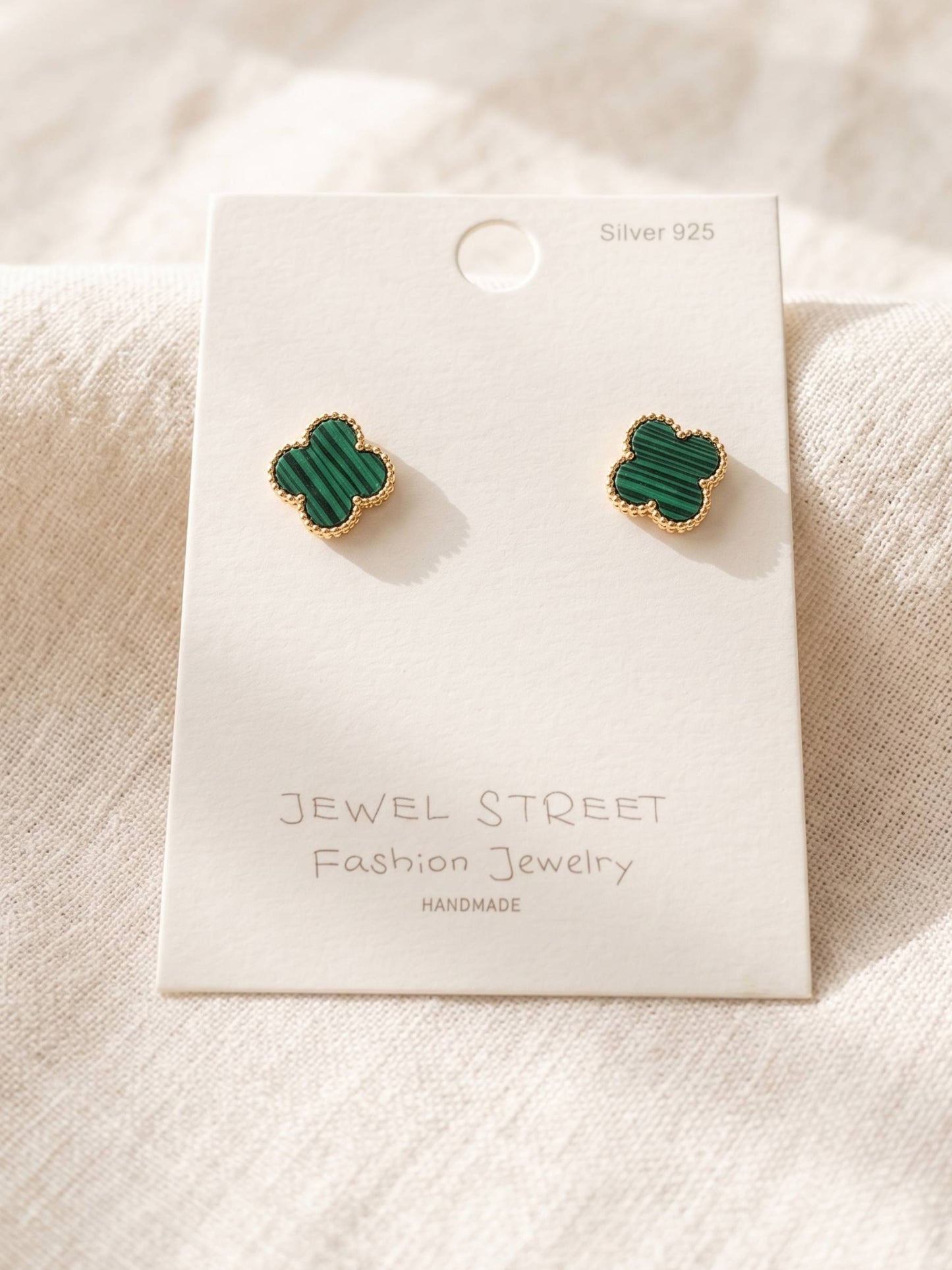 Green clover-shaped earrings on a 'Jewel Street' branded card against a beige fabric background.