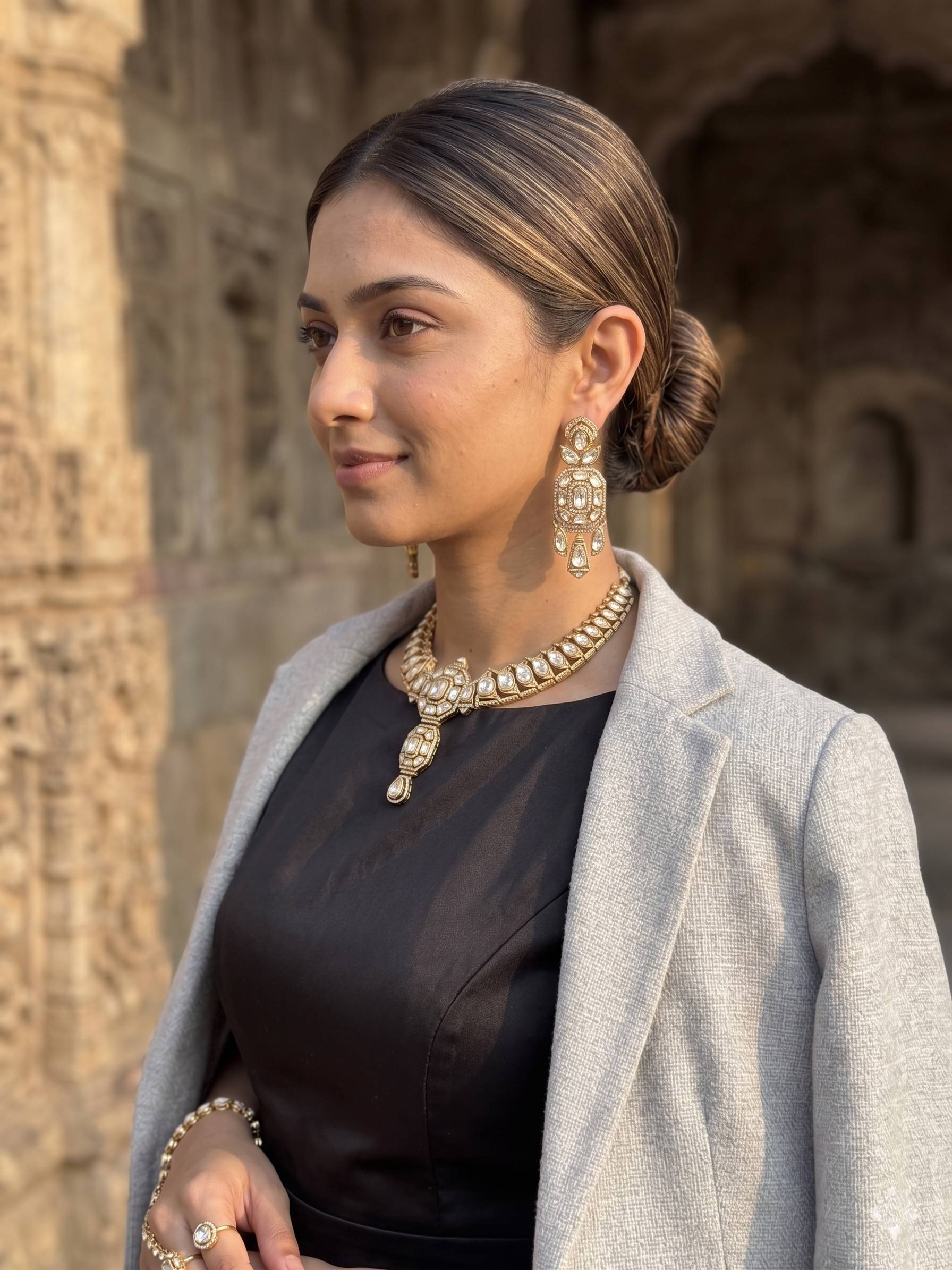 Woman wearing a black outfit with a gray coat and gold jewelry, standing in front of an architectural background.
