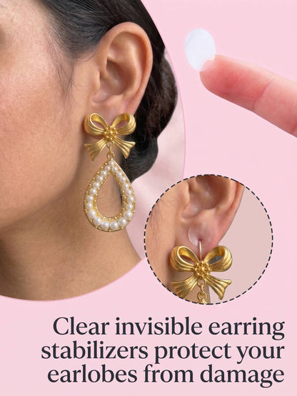 Gold bow-shaped earrings with pearl details worn by a person, on a pink background.