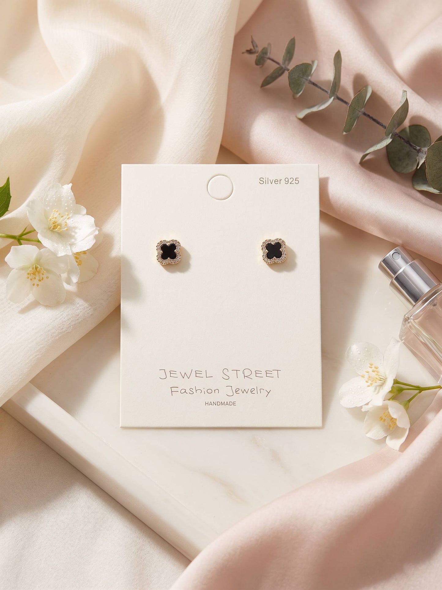 Pair of earrings on a 'Jewel Street' card with flowers and a perfume bottle in the background