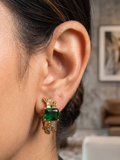 Close-up of an ear wearing a gold earring with a green gemstone, blurred indoor background