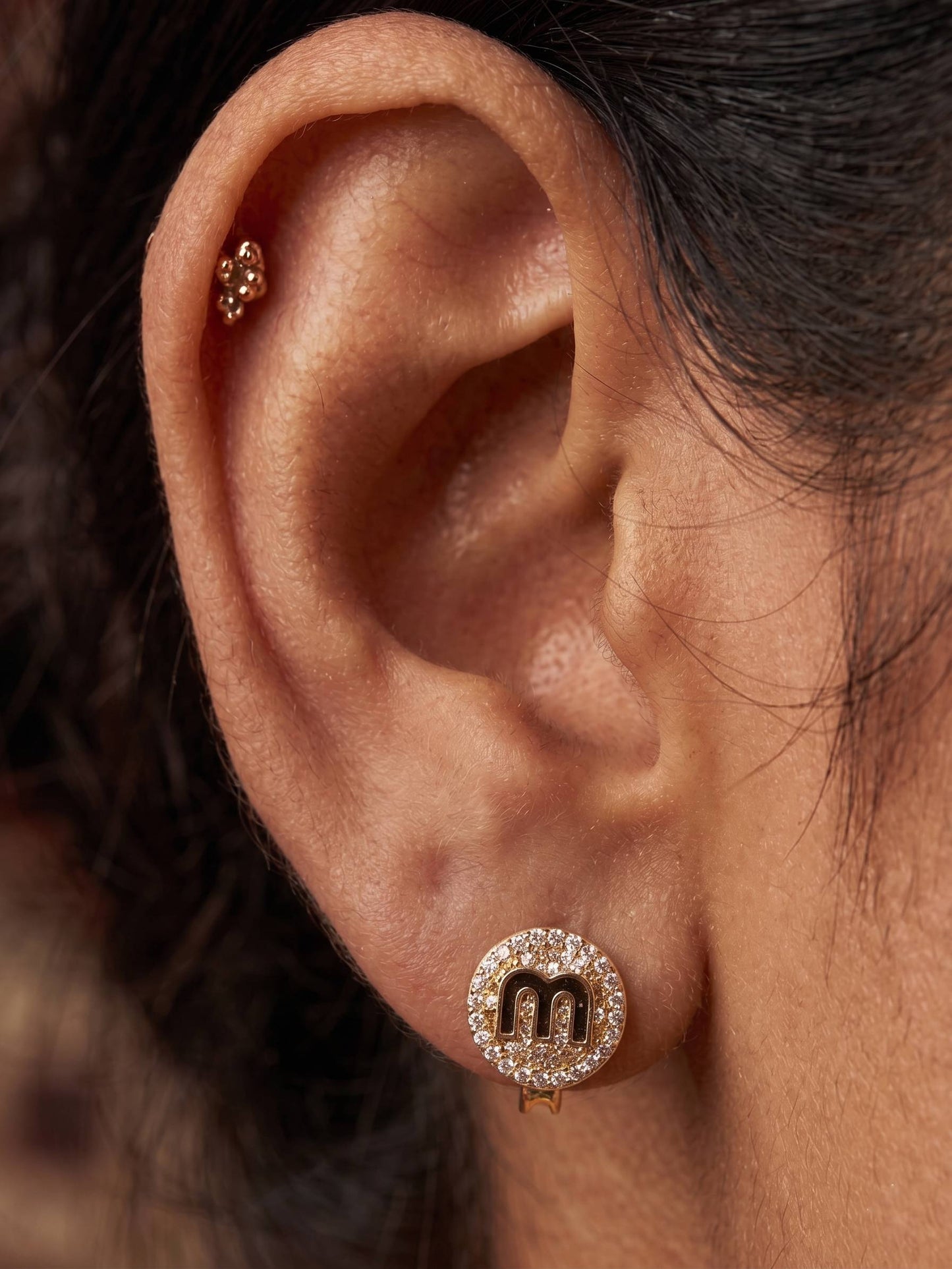 Close-up of an ear wearing a gold earring with a letter 'M'.
