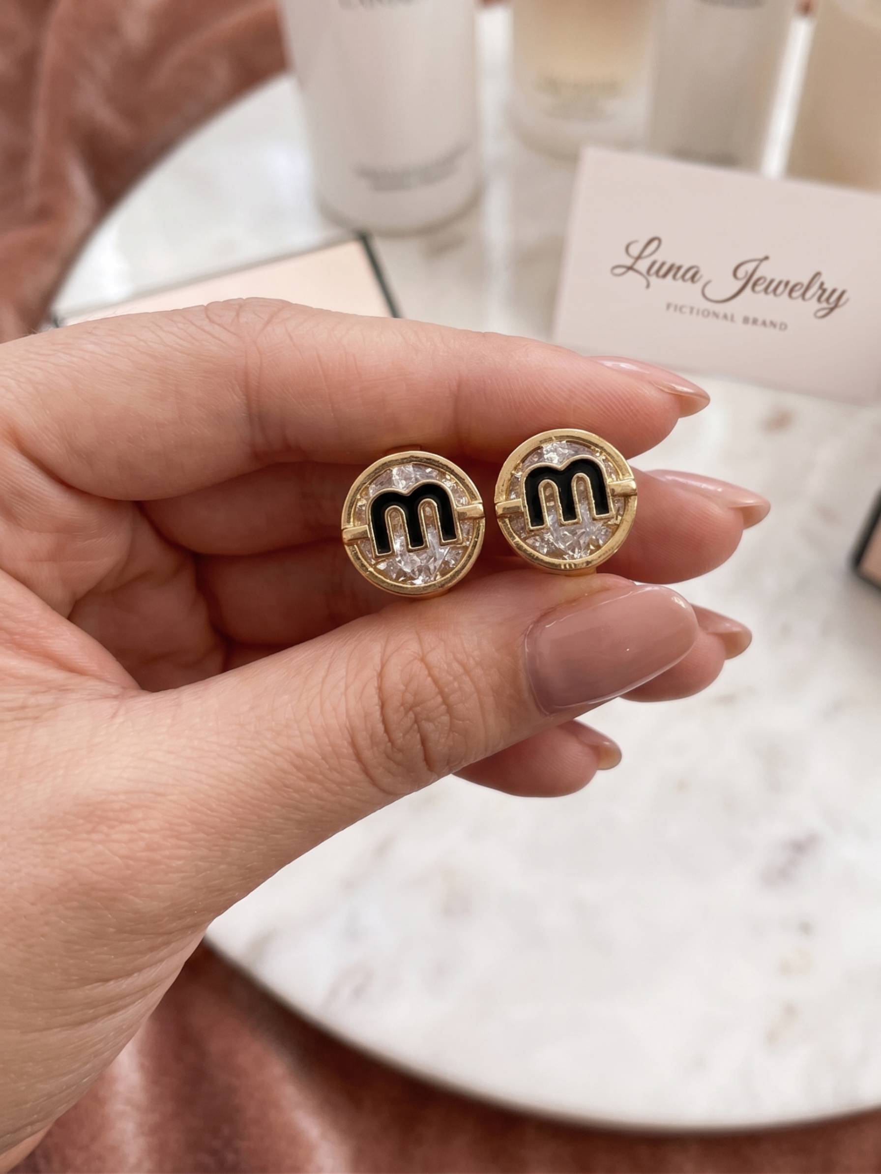 Gold earrings with 'm' design held by a hand, branded 'Luna Jewelry'.