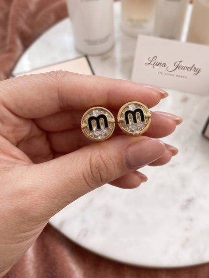 Gold earrings with 'm' design held by a hand, branded 'Luna Jewelry'.