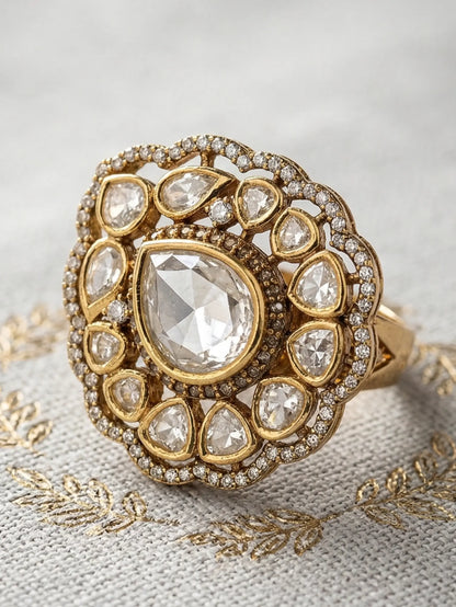 Gold ring with a large central diamond and smaller diamonds in a decorative setting on a textured fabric background.