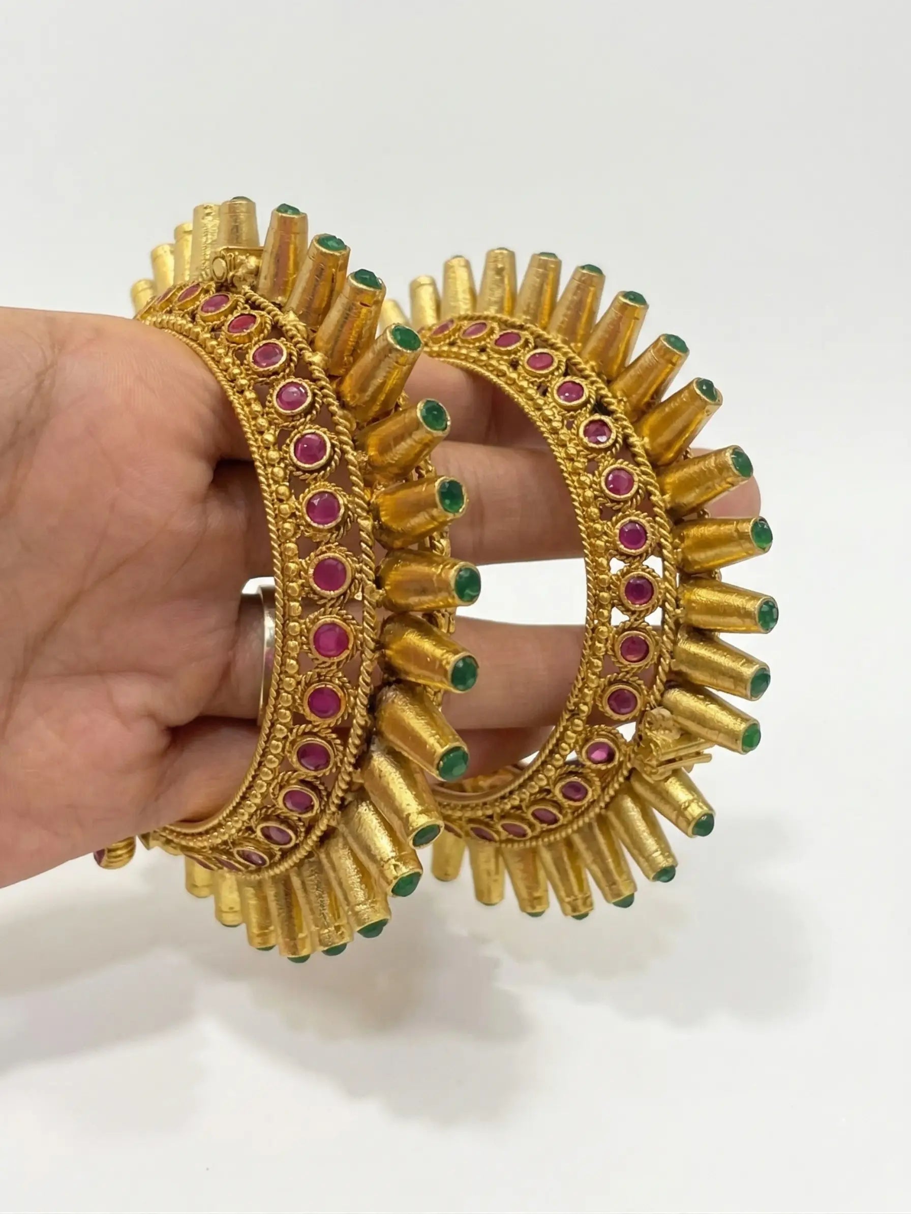 Gold earrings with red and green stones held by a hand on a white background