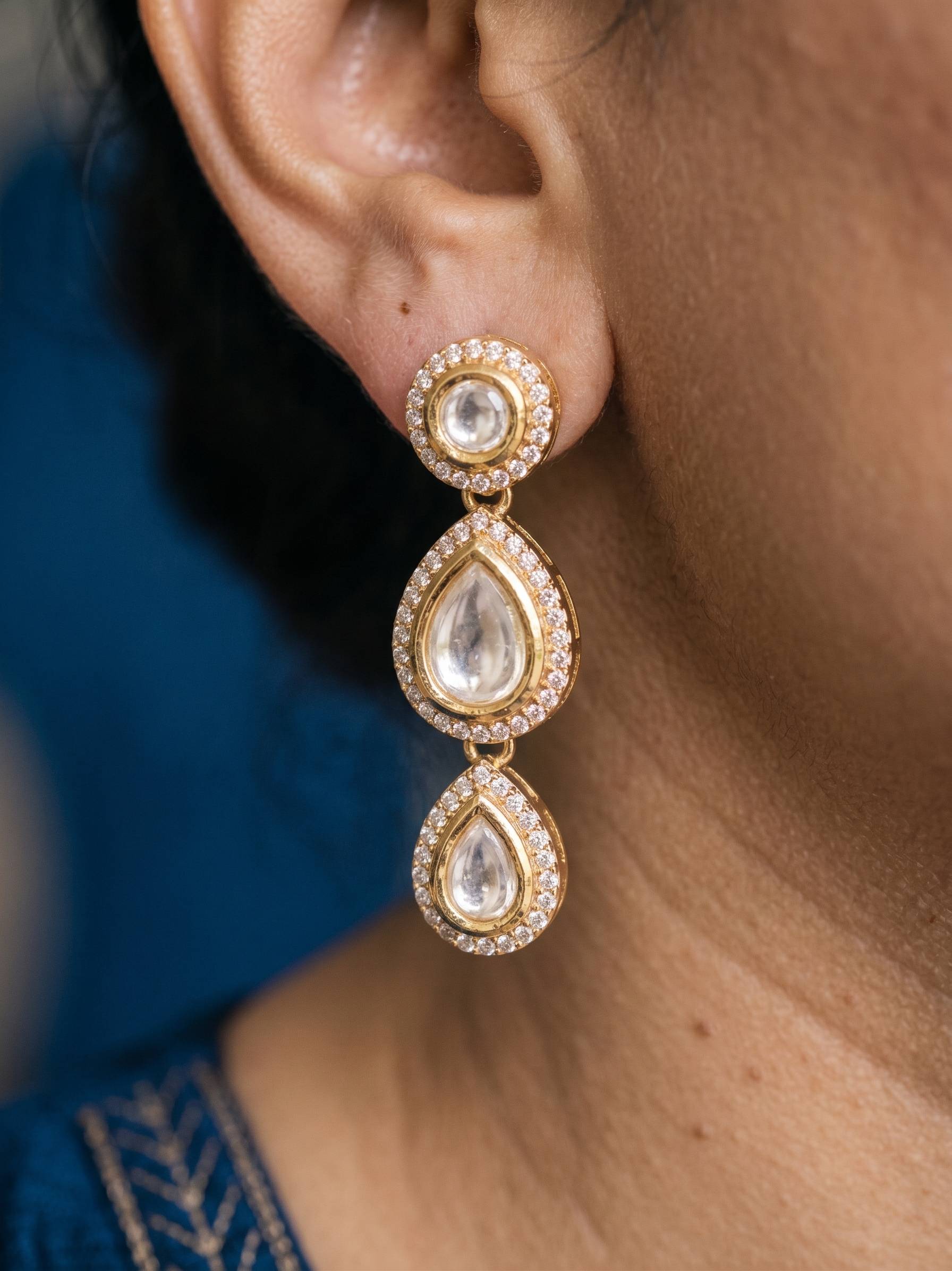 Gold earrings with clear gemstones worn by a person against a blurred background